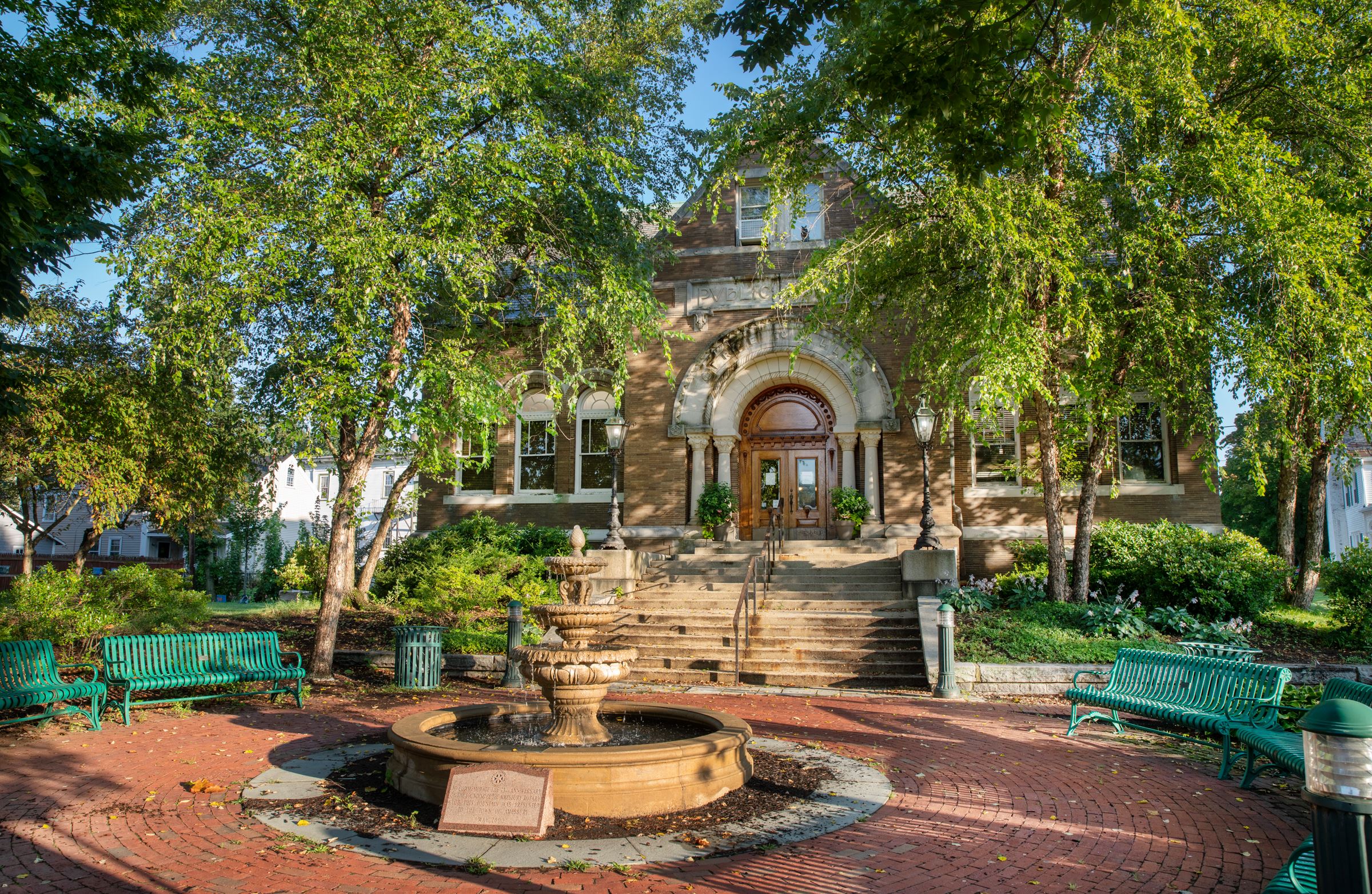 view of the plaza showing some of the public benches (green metal) and the fountain, facing the library's front steps and front doors