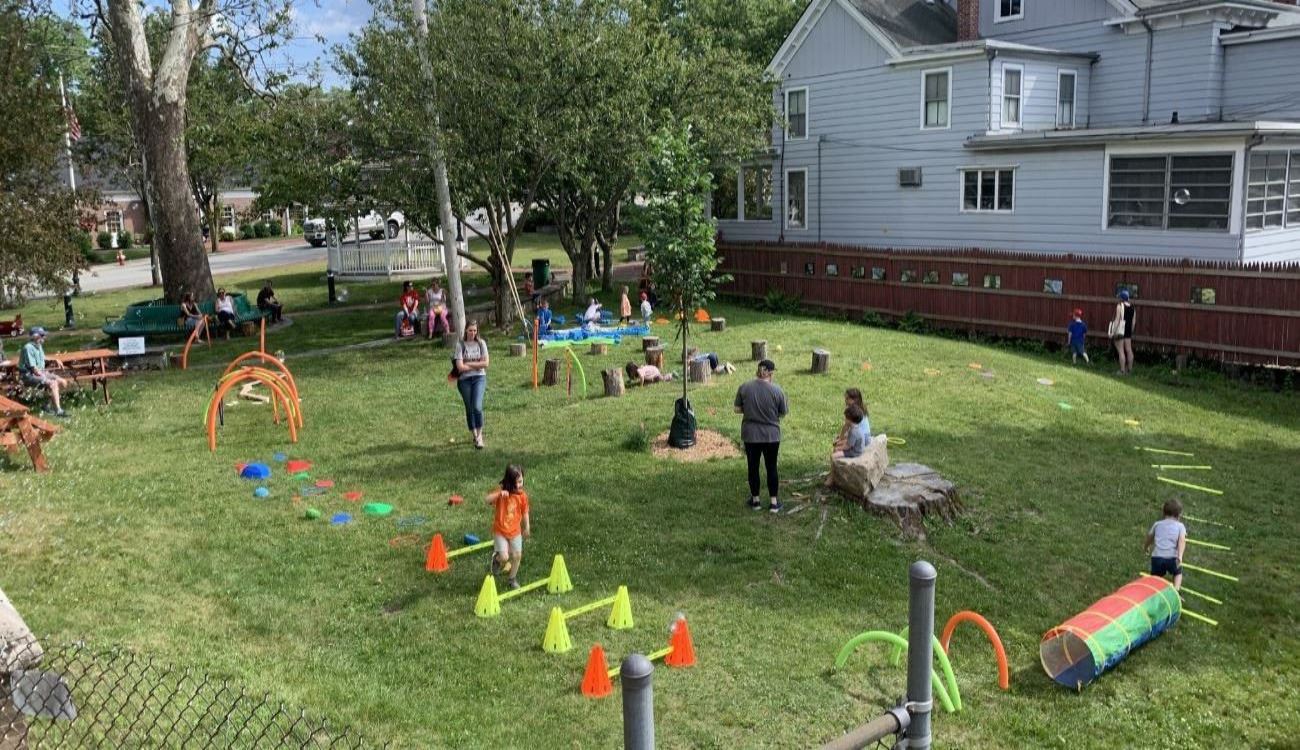 view of the library's side lawn facing Main St, showing games during a children's event in the foreground and a storywalk along the fence in the background